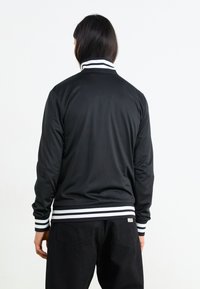 Sort jakke med høj krave, forsynet med hvide striber på kraven og manchetterne. Fremstillet af glat, letvægtsstof.