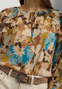Květnová blůza s řasenými rukávy, převážně hnědé, modré a žluté barvě, s mašlí na výstřihu a texturovanou tkaninou.