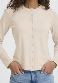 Bež dolg rokav kardigan z okroglim izrezom, zapenjanjem na gumbe in rebrasto teksturo. Oblečen s svetlo modrimi kavbojkami.