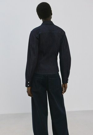 Persona con capelli corti che indossa una giacca di jeans scura aderente e pantaloni di jeans scuri larghi, in piedi di fronte a una parete chiara e liscia.