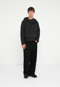 Schwarze Jacke mit Kapuze, leichtem Stoff, Reißverschluss und Seitentaschen; kombiniert mit locker sitzenden schwarzen Hosen und Schuhen.