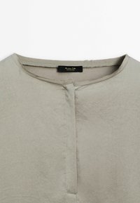 Blouse gris clair avec un col rond, une patte de boutonnage sur le devant, une texture de tissu lisse et un léger éclat. Étiquette visible au niveau de l'encolure.