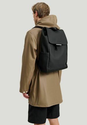 Black backpack with a flap closure, made of smooth material, featuring side pockets and adjustable straps, worn over a brown rain jacket.