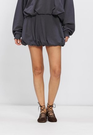 Femme portant une mini jupe à ourlet bouffant gris foncé, un haut à manches longues assorti et des chaussures marron plates à lacets, debout devant un fond uni.