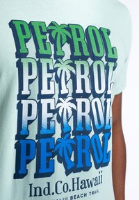 Světle zelené bavlněné tričko s velkým modrým a zeleným nápisem "PETROL", doplněným grafikou palmami, vytištěným v naskládaném designu.