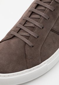 Sneaker in suede marrone con punta arrotondata, lacci piatti e suola in gomma bianca con dettagli texturizzati.