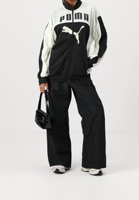 Schwarz-weiße Sportjacke mit PUMA-Logo, kombiniert mit schwarzen weit geschnittenen Hosen und einer kleinen glänzenden schwarzen Handtasche. Sneakers mit hellen Akzenten.