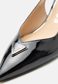 Escarpin noir verni à bout pointu avec un logo triangulaire en métal Guess USA sur le devant.