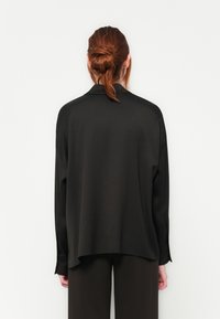 Svart langermet bluse laget av glatt stoff, med en avslappet passform og lett buet kant, sett bakfra.