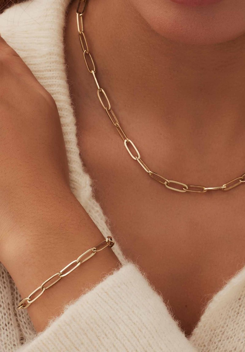 Le collier et le bracelet en chaîne dorée présentent des maillons ovales allongés. Les deux articles ont une finition lisse et polie et sont portés sur un pull crème.