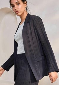 Námořnický strakatý blazer s přiléhavým střihem a ostrými klopami, vrstvený přes světle šedý žebrovaný top s leskem a jemnou légou.