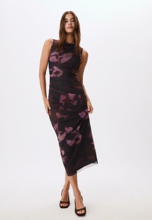 Woman standing against white background wearing a sleeveless black and purple floral-patterned midi dress and black high-heeled sandals.