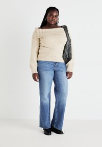 Sieviete ar garām bizēm, valkājot plecus atsedzošu bēša krāsas džemperi, zilas džinsas, melnus apavus un tumšu šalli ar rakstu pār vienu plecu.