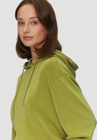 Haut à capuche vert avec une coupe décontractée, capuche à cordon, texture douce et poignets côtelés. Il comprend une poche kangourou et des détails de couture minimaux.