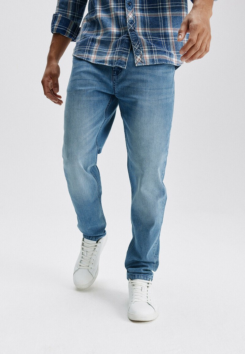 Jasnoniebieskie dżinsy o luźnym kroju, z subtelnymi przetarciami i bocznymi kieszeniami, w połączeniu z białymi sneakersami i niebieską koszulą w kratę.