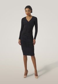 Svart långärmad klänning med draperade detaljer, med V-ringning och kroppsnära siluett, i kombination med nude högklackade skor.