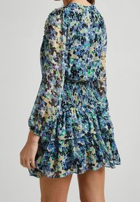 Kobieta ubrana w sukienkę w niebiesko-zielono-czarny kwiatowy wzór, z długimi rękawami i gumowanym pasem, pokazana z tyłu.