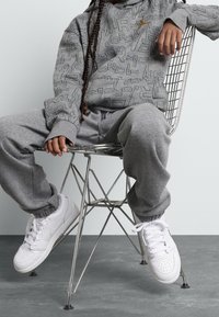 Grauer Hoodie und Jogginghose mit wiederholtem Textmuster, kombiniert mit weißen Nike-Sneakern, sitzend auf einem Drahtstuhl vor einem hellen Hintergrund.