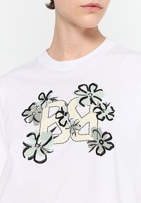 Bílý bavlněný tričko s velkým potiskem s nápisem "BS" v žluté barvě, obklopené stylizovanými černými a zelenými květinami a listy.