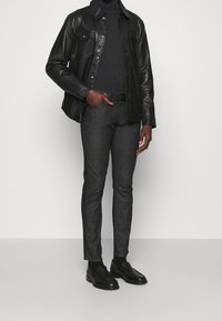 Veste-chemise en cuir noir portée sur un col roulé gris foncé, associée à un jean slim foncé et des chaussures noires sans lacets, le tout dans un style minimaliste.