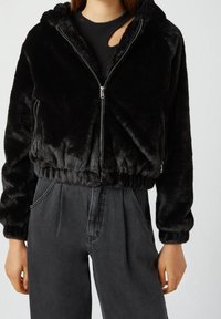 Veste courte en fausse fourrure noire avec fermeture éclair, capuche et ourlet élastiqué. Associée à un pantalon en denim foncé taille haute.