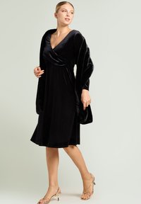 Schwarzes Samtwickelkleid mit V-Ausschnitt, gerafftem Oberteil und ausgestelltem Rockteil. Dazu ein passender Kunstpelz-Wickelmantel. Goldene Riemchenabsätze.