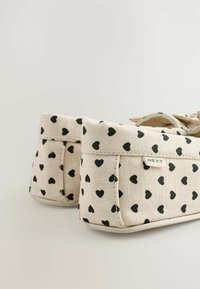 Beige canvas slip-on kengät, joissa on pieniä mustia sydämiä kuvastava kuvio. Varustettu taitetulla resorilla ja ommellulla tarralla, jossa on brändin nimi.