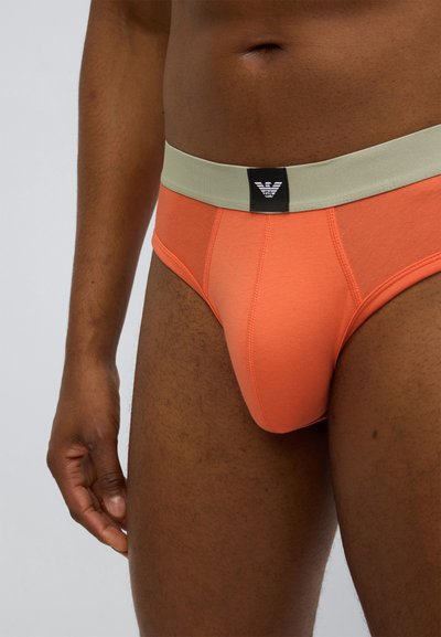 Orange bomullsbriefs med en ljusgrå midjeresår med en svart logotyp. Tyget har en slät textur och en åtsittande design.