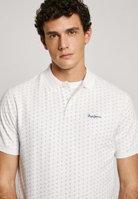 White polo shirt featuring a small blue anchor pattern, two-button placket, and contrasting blue logo embroidery on the left chest.