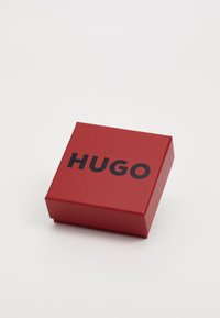 Raudonas kvadratinis dėžutė su lygiu paviršiumi, viršuje esantis ryškus juodas tekstas "HUGO". Nėra jokių papildomų raštų ar akcentų.