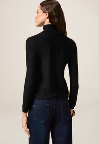 Pull à col roulé noir au design ajusté, avec une texture mouchetée. Associé avec un jean large bleu foncé.