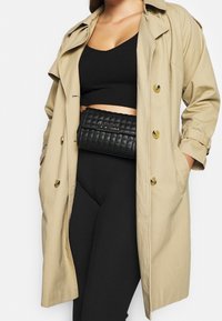 Beżowy płaszcz trencz o swobodnym kroju, z dużymi przednimi kieszeniami i złotymi guzikami, noszony na czarnym prążkowanym krótkim topie i dopasowanych legginsach. Czarna pikowana torebka na pasek.