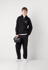 Black zip-up sweatshirt with CK logo, paired with black pants and white sneakers. Model holds a small black handbag.