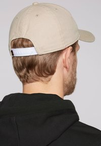 Khaki cap made of fabric with a curved brim, featuring stitched detailing and an adjustable white plastic strap at the back. Black hoodie visible.