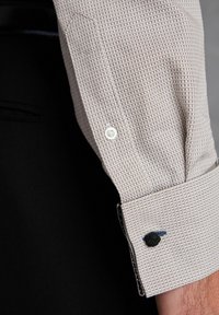 Woven shirt w kolorze jasnego beżu o teksturowanej tkaninie, z białym guzikiem i czarnym haftowanym akcentem na mankiecie.