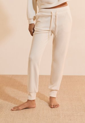Personne portant un pantalon de jogging en maille côtelée couleur crème avec des poignets aux chevilles et un haut court assorti, debout pieds nus sur un tapis beige.