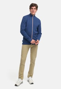 Blå zip-jakke med teksturert stoff, oransje glidelåsdetalj, kombinert med beige bukser og hvite sneakers mot en nøytral bakgrunn.