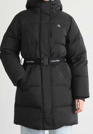 Zwarte gewatteerde Calvin Klein jas met capuchon, ritssluiting en geborduurde riem, gedragen door een persoon met handen in zakken tegen een witte achtergrond.