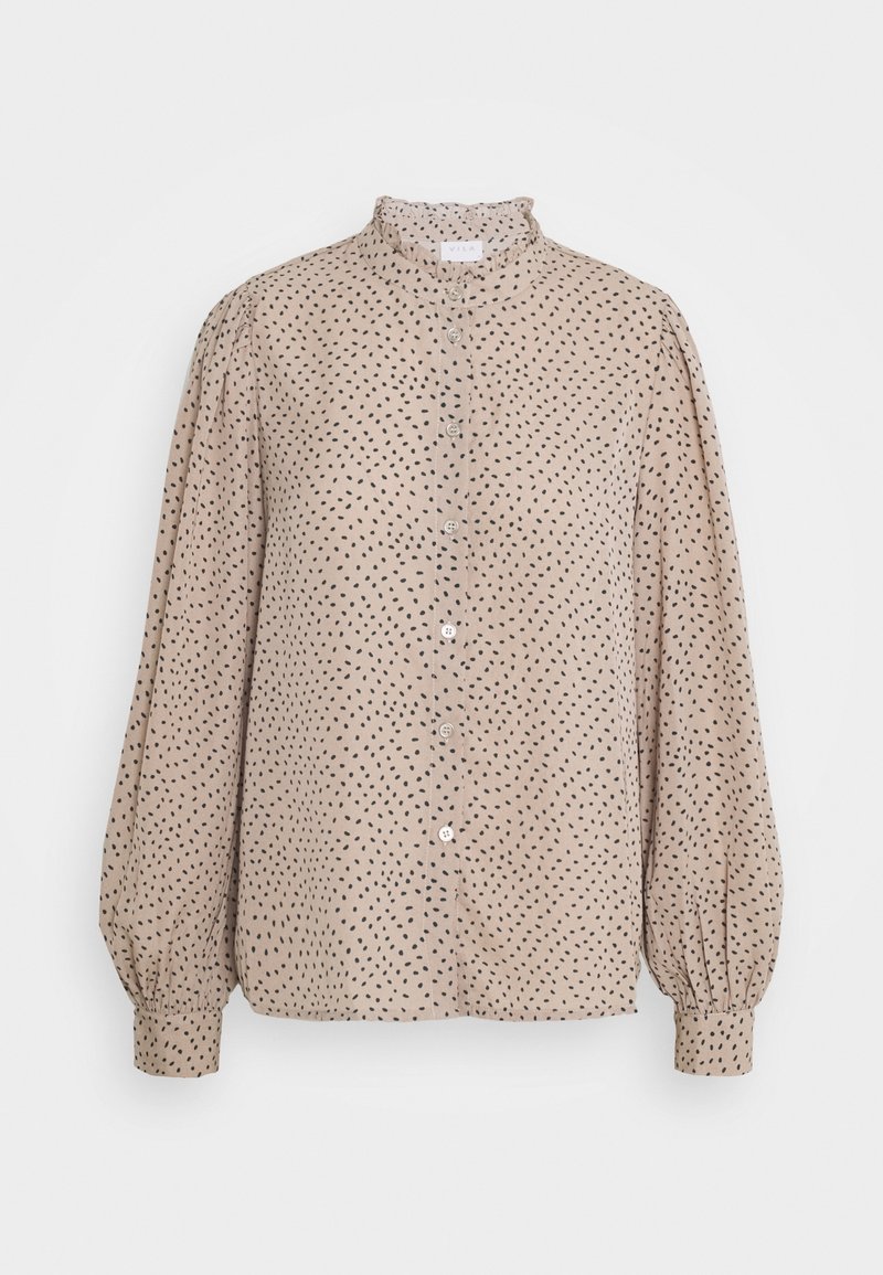 Blouse beige à manches longues avec un motif de points noirs irréguliers, fermeture boutonnée sur le devant, col haut à volants et poignets froncés.