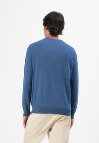 Uomo con capelli scuri che indossa un maglione blu a maniche lunghe e pantaloni beige, in piedi di spalle su uno sfondo bianco semplice.