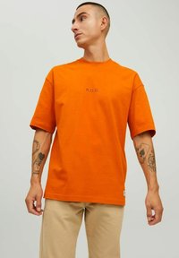 Oranžs kokvilnas t-krekls ar īsām piedurknēm un brīvu piegriezumu, kas priekšpusē rotāts ar melnu tekstu "R.D.D.".