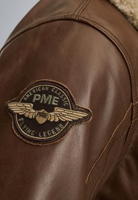 Smešak kožni jakna s krznom ovratnikom; sadrži okrugli pečat s krilima, "PME" i tekst "Flying Legend". Teksturirani, robusni materijal.