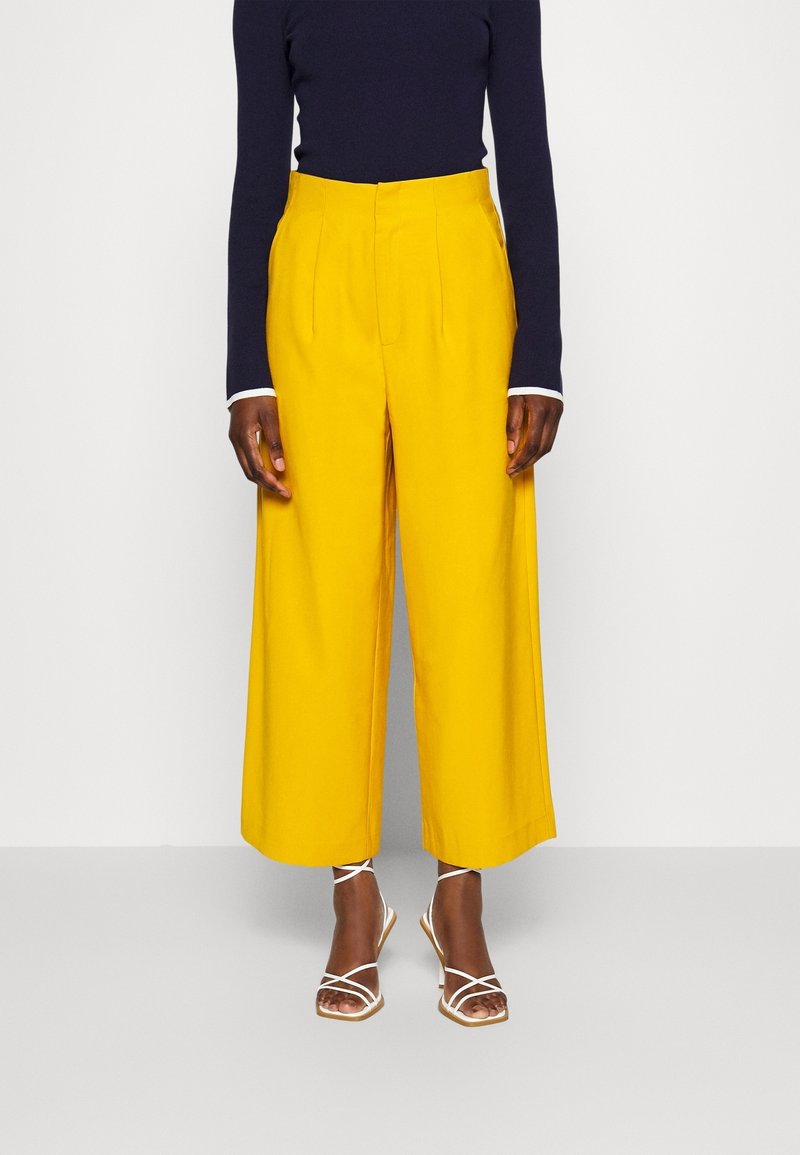 Personne portant un pantalon jaune taille haute à jambes larges, un haut bleu marine à manches longues et des sandales blanches à talons et à brides, debout devant un fond uni.