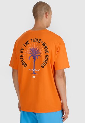 Mężczyzna zwrócony tyłem, ubrany w pomarańczowy t-shirt z niebieską palmą i napisem „Napędzani przez przypływy, jeźdźcy fal” na plecach.