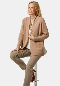 Camel kleurige sjaal cardigan met een zachte, gestructureerde afwerking, met een open voorkant, geribbelde manchetten en een pocket aan de linkerkant, gecombineerd met beige broeken.