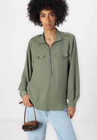 Olivenfarget bluse med halvlukking foran, to brystlommer og knappede mansjetter, laget av glatt stoff. Modellen holder en liten brun håndveske.