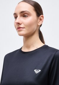 Femme aux cheveux bruns attachés, portant des boucles d'oreilles créoles dorées et une chemise noire avec un petit logo blanc en forme de cœur sur la poitrine.