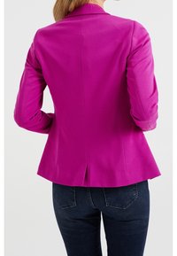 Blazer ajusté en tissu rose vif, avec une fente simple à l'arrière, des revers structurés et des manches longues. Porté avec un jean noir.