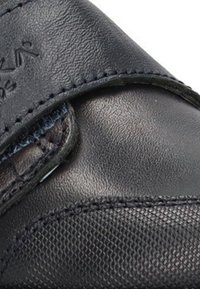 Zapato de cuero negro con superficie texturizada, que presenta una correa de velcro. Muestra un acabado suave y costuras duraderas a lo largo de los bordes.