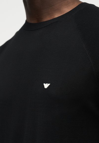 Κοντινό πλάνο ενός ατόμου που φοράει μαύρο μπλουζάκι με στρογγυλή λαιμόκοψη και μικρό λευκό λογότυπο στο στήθος.